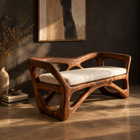 Bench with a sculpted open wooden frame, low arms and a pale cushion, three-quarter view showing the grain