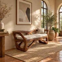 Bench with a sculpted open wooden frame, low arms and a pale cushion in a bright entryway with arched windows and plants
