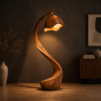 Floor lamp with a long arching wooden stem and a carved hood shade in a dark studio against a charcoal plaster wall