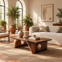 Thick slab coffee table on carved arch legs with circular cutouts in a bright living room with arched windows and plants