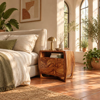 Barrel sided nightstand with an open top cubby and a swirled grain front in a bright bedroom with arched windows and plants