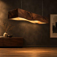 Long beam pendant light with a wavy lower edge and a warm glow in a dark studio against a charcoal plaster wall