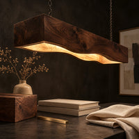 Corner detail of long beam pendant light with a wavy lower edge and a warm glow beside wood, brass and linen samples