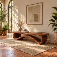Low wood bench with a wavy slab top on an open ribbon loop base in a bright entryway with arched windows and plants
