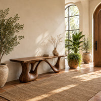 Long low slab bench with a wavy edge on a sculpted base in a bright entryway with arched windows and plants
