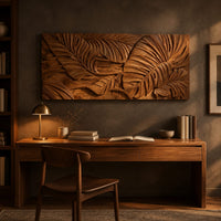 Carved wood wall art panel with overlapping tropical leaves in deep relief above a desk in a dark study corner