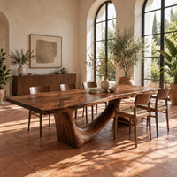 Dining table with a flowing slab top on a branching sculpted base in a bright dining room with arched windows and plants