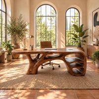 Sculpted wooden desk with branching legs and open shelves at one end in a bright home office with arched windows and plants