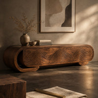 Corner detail of capsule shaped tv stand with carved wave panels and a scroll end ledge beside wood, brass and linen samples