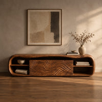 Corner detail of low capsule shaped wooden tv stand with wave carved panels and open end shelves beside wood, brass and linen samples