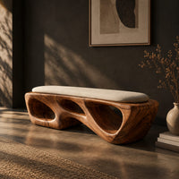 Chunky wooden bench with sculpted cutouts and a slim cream cushion on top, three-quarter view showing the grain