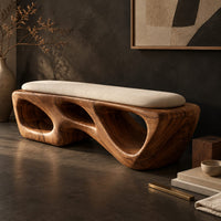 Corner detail of chunky wooden bench with sculpted cutouts and a slim cream cushion on top beside wood, brass and linen samples