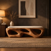 Chunky wooden bench with sculpted cutouts and a slim cream cushion on top in a dark entryway at golden hour