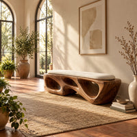 Chunky wooden bench with sculpted cutouts and a slim cream cushion on top in a bright entryway with arched windows and plants