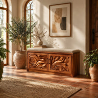 Shoe bench with two carved relief doors and a padded seat on top in a bright entryway with arched windows and plants