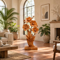 Carved wood flower sculpture with open blossoms and leaves on a block base in a bright living room with arched windows and plants