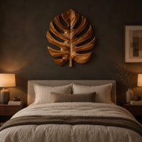 Monstera leaf carved from wood with deep slits and a short stem above a bed in a dusk lit bedroom