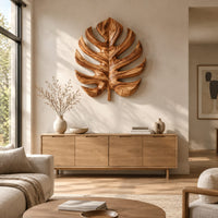 Monstera leaf carved from wood with deep slits and a short stem above a light oak sideboard in daylight