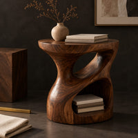 Corner detail of carved pedestal side table with a twisting opening and a low open nook beside wood, brass and linen samples