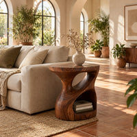 Carved pedestal side table with a twisting opening and a low open nook in a bright living room with arched windows and plants