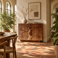 Two door sideboard with a carved tree branch relief across the fronts in a bright dining room with arched windows and plants