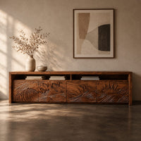 Corner detail of long tv stand with four carved doors showing ocean waves and a rising sun beside wood, brass and linen samples