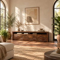 Long tv stand with four carved doors showing ocean waves and a rising sun in a bright living room with arched windows and plants