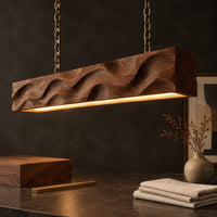 Corner detail of long wood pendant light with carved waves and a warm glow along its underside beside wood, brass and linen samples