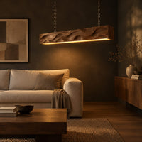 Long wood pendant light with carved waves and a warm glow along its underside in a dark living room at golden hour