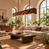 Long wood pendant light with carved waves and a warm glow along its underside in a bright living room with arched windows and plants