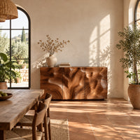 Sideboard with three flush doors and a rolling carved wave relief front in a bright dining room with arched windows and plants