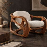 Corner detail of chair with a chunky sculpted frame, large oval cutouts and a pillow back beside wood, brass and linen samples