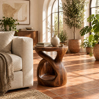 Chunky side table with a looping open body and thick flat top in a bright living room with arched windows and plants