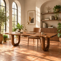 Desk with a live edge slab top and two thick sculpted legs in a bright home office with arched windows and plants