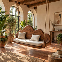 Cloud shaped swing with a carved wavy backrest and cream seat cushion in a bright porch with arched windows and plants