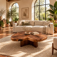 Clover shaped coffee table with a thick top and cross shaped pedestal in a bright living room with arched windows and plants