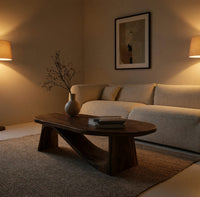 Coffee table on angled plank legs with a swooping brace in an evening room lit by two lamps