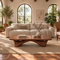 Low coffee table with a carved organic base and a soft edged top in a bright living room with arched windows and plants