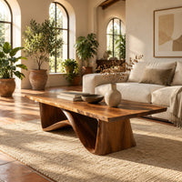 Coffee table with a tapered block pedestal and a swooping wood loop in a bright living room with arched windows and plants