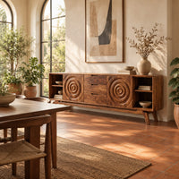 Sideboard with ring carved panels and open end shelves on angled legs in a bright dining room with arched windows and plants