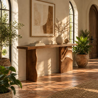 Console table with a slab top and one leg carved in flowing waves in a bright entryway with arched windows and plants