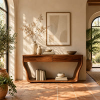 Console table with flared sculpted sides, a low shelf and two drawers in a bright entryway with arched windows and plants