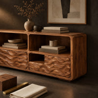 Corner detail of low console cabinet with three open cubbies above two wave carved front panels beside wood, brass and linen samples