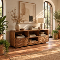 Low console cabinet with three open cubbies above two wave carved front panels in a bright entryway with arched windows and plants