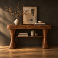Console table with two waisted sculpted legs and a single open shelf below in a dark studio against a charcoal plaster wall