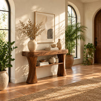 Console table with two waisted sculpted legs and a single open shelf below in a bright entryway with arched windows and plants