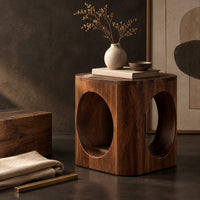 Corner detail of cube shaped side table with large oval cut outs and softened edges beside wood, brass and linen samples
