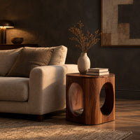 Cube shaped side table with large oval cut outs and softened edges in a dark living room at golden hour