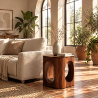 Cube shaped side table with large oval cut outs and softened edges in a bright living room with arched windows and plants