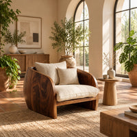 Barrel chair with curved solid wood sides and a deep cream seat cushion in a bright living room with arched windows and plants